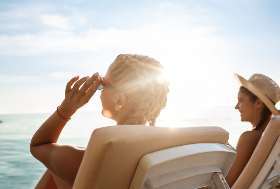 Atrás da mulher loira de cabelos entrançados, que ajusta os óculos de sol enquanto relaxa numa espreguiçadeira à beira-mar, vê-se outra mulher a sorrir sob a luz forte do sol poente.
