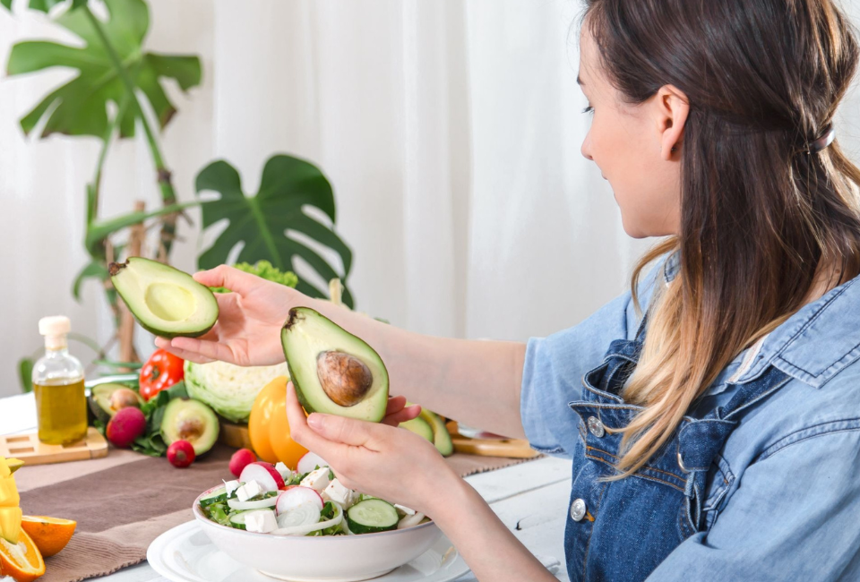Mulher sentada à mesa com um prato de salada à frente e a pegar num abacate cortado ao meio