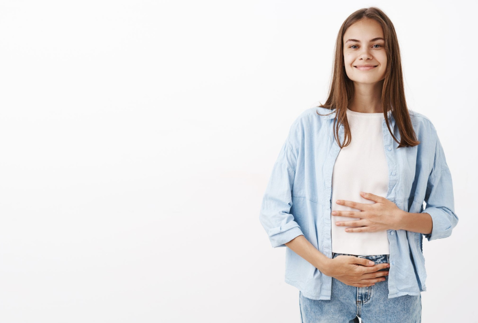 Mulher jovem virada de frente, com as duas mãos na barriga e com um sorriso que demonstra bem-estar