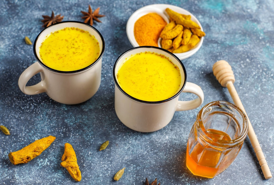 Duas chávenas de golden milk junto de raízes de curcuma e copo de mel