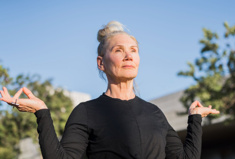 Uma mulher sénior, com o cabelo grisalho apanhado num coque, pratica meditação ao ar livre com as mãos em mudra e o rosto voltado para o sol.