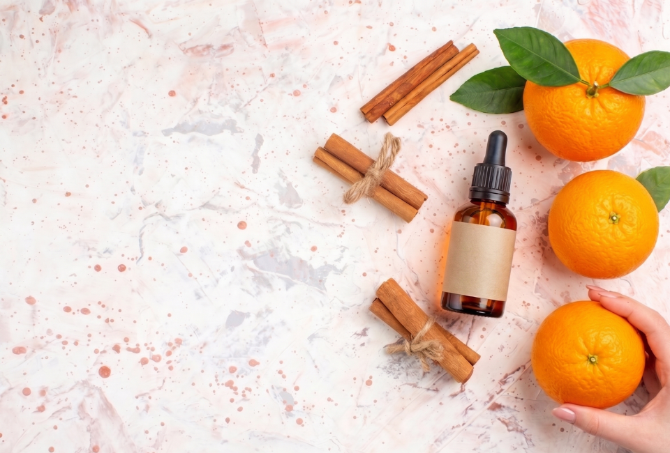 Vista superior de um frasco conta-gotas de óleo essencial de laranja sobre mármore, cercado por laranjas inteiras e paus de canela, com uma mão a segurar uma laranja