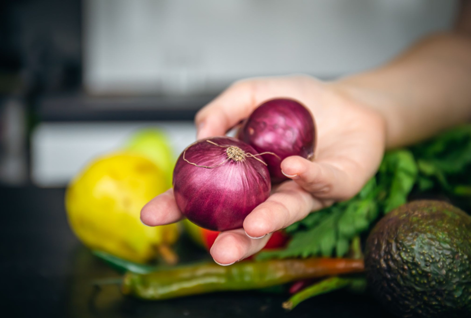 Mão a segurar duas cebolas roxas pequenas, com outros vegetais e frutas frescos ao fundo, como peras amarelas, pimenta verde e um abacate.