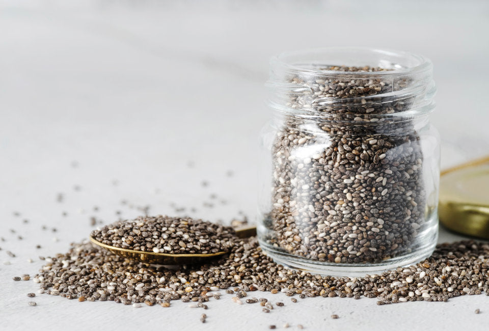Frasco de sementes de chia em cima de mesa branca com sementes espalhadas à volta