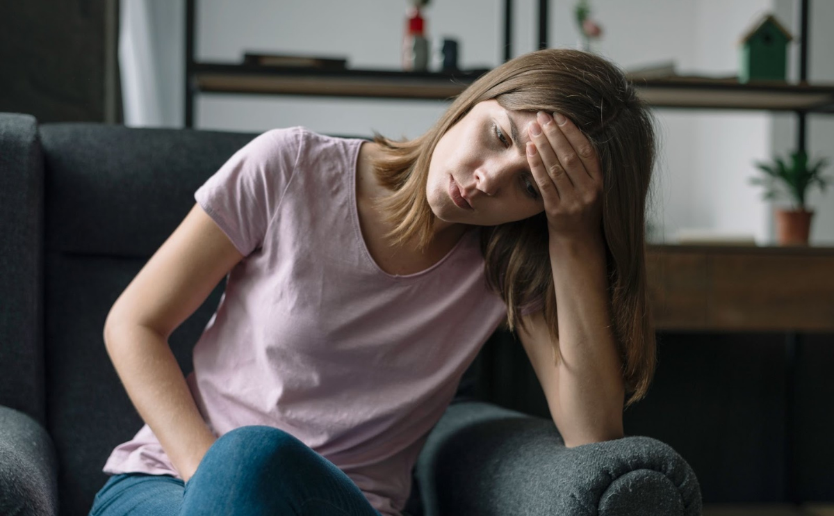 Mulher sentada no sofá com uma mão na testa e uma na barriga com sintomas da SIBO